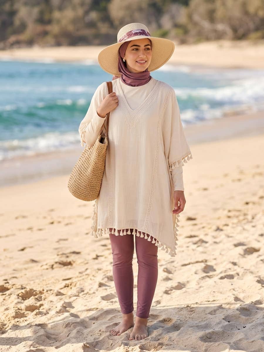 Femme portant un maillot pudique sous un caftan de plage crème fluide, un chapeau posé par-dessus le hijab